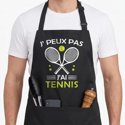 Tablier de cuisine noir avec deux raquettes de tennis blanches croisées et une balle jaune vif au centre du design imprimé.