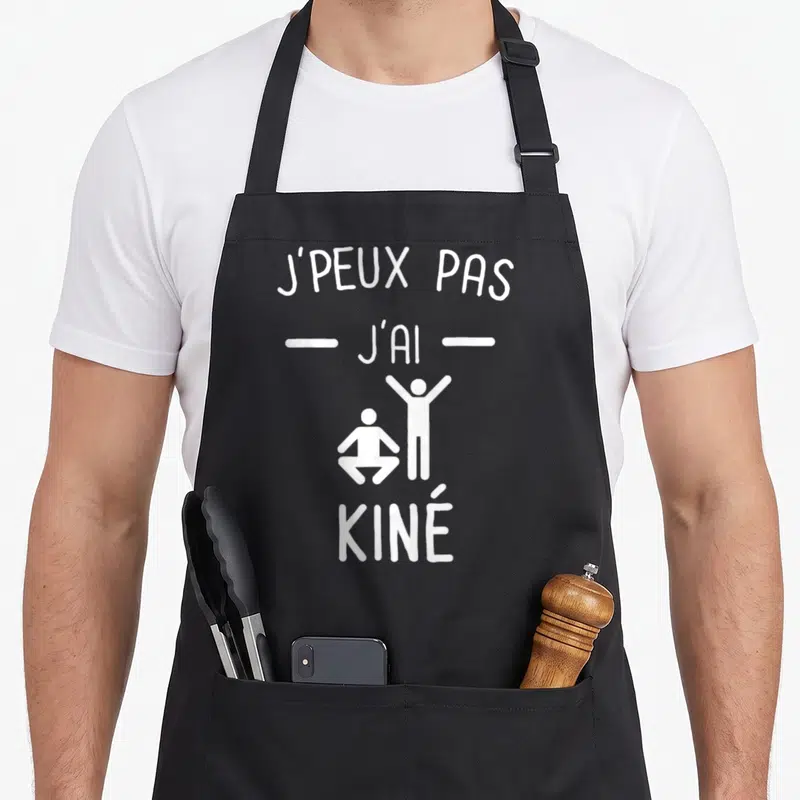 Tablier de cuisine noir avec inscription blanche J'peux pas j'ai Kiné et deux silhouettes stylisées.