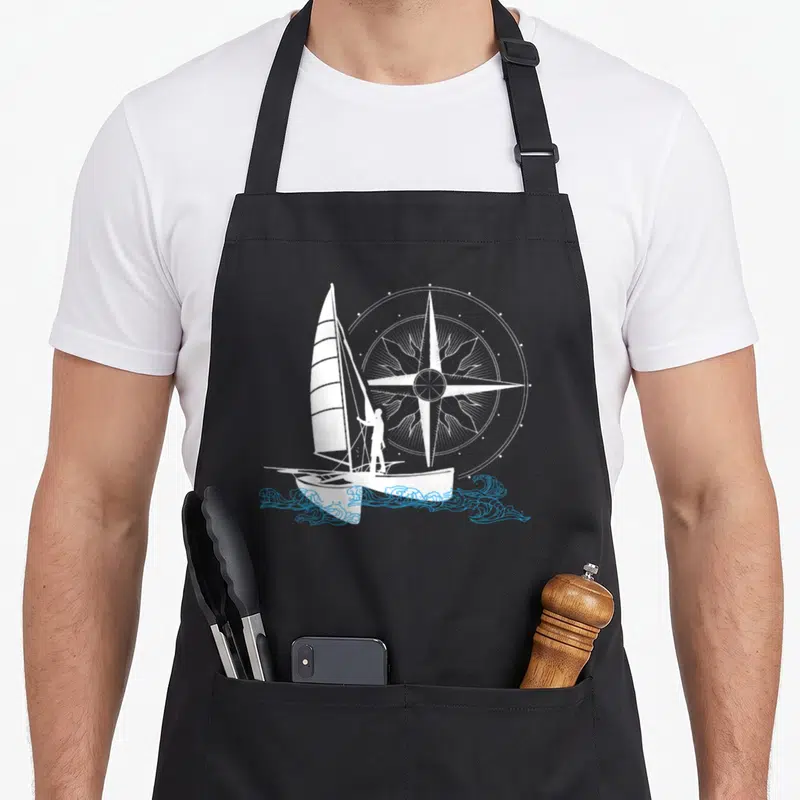 Tablier de cuisine noir avec voilier blanc, silhouette de marin et grande boussole sur fond bleu vif.