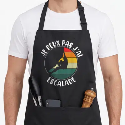 Tablier de cuisine noir avec motif circulaire montrant un silhouetté de grimpeur sur fond rayé multicolore et texte blanc.