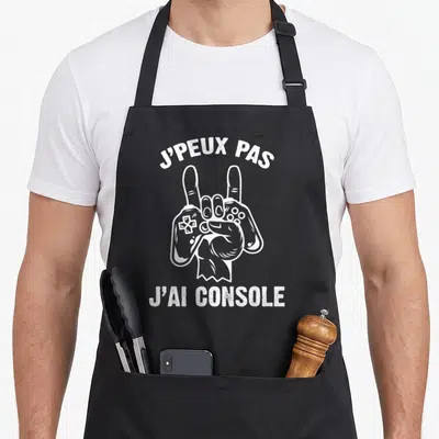 Tablier de cuisine noir avec main blanche en signe de rock tenant une manette de jeu vidéo et texte imprimé.