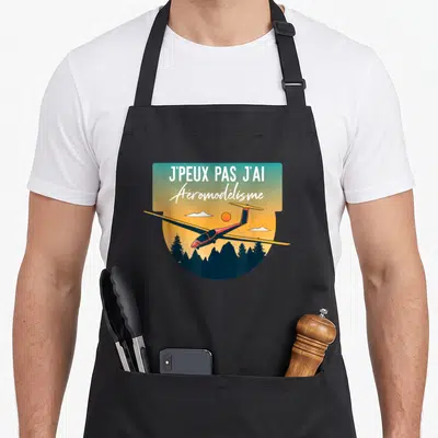 Tablier de cuisine noir avec illustration d'un planeur rouge volant au-dessus de montagnes et d'arbres sous un ciel aux couleurs dégradées.