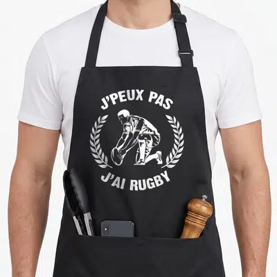 Tablier de cuisine noir avec un joueur de rugby posant le ballon et le texte J'peux pas j'ai rugby en blanc.