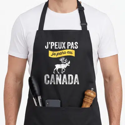 Tablier de cuisine noir avec l'inscription j'peux pas je pars au Canada et un dessin de cerf blanc sur fond jaune.