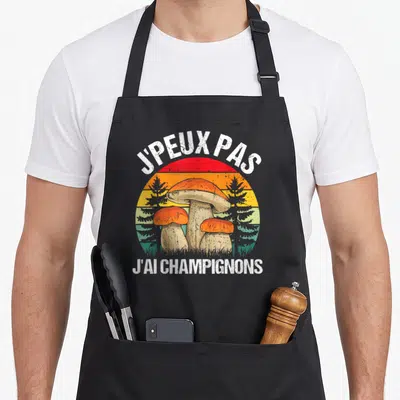 Tablier de cuisine noir avec illustration rétro de trois cèpes orange devant des sapins sur fond arc-en-ciel délavé.
