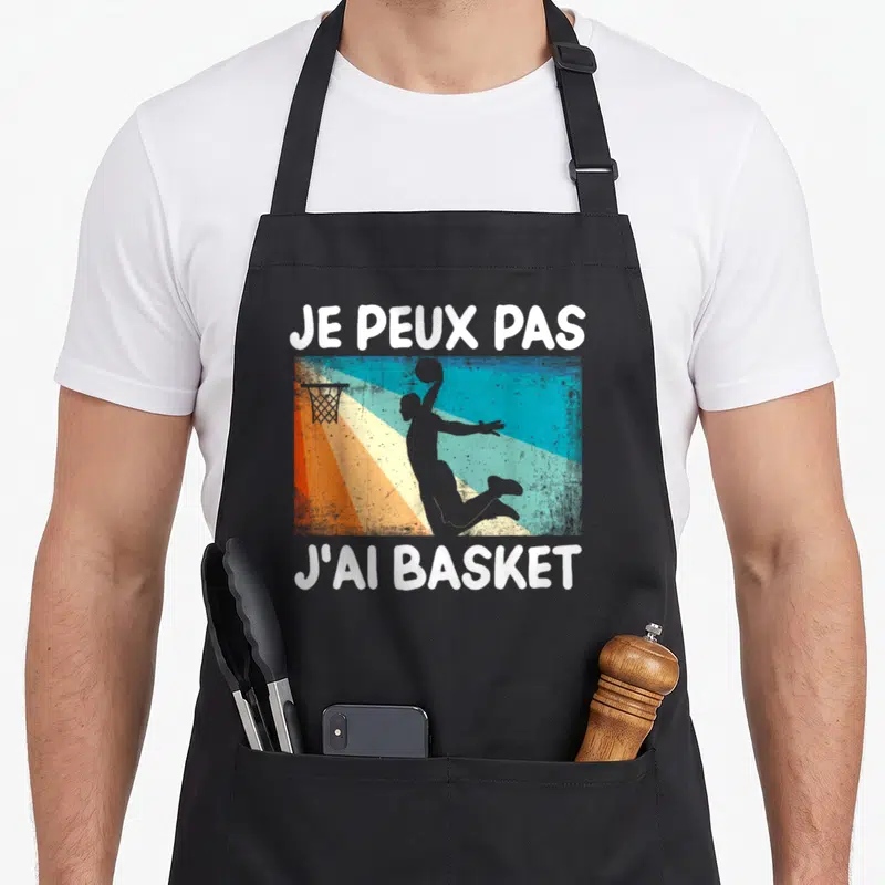 Tablier de cuisine noir avec silhouette de joueur de basket et texte je peux pas j'ai basket en blanc sur fond rayé coloré.