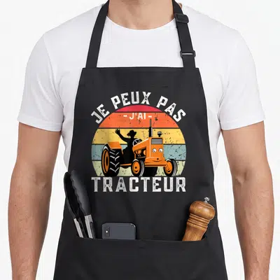 Tablier de cuisine noir avec illustration vintage d'un tracteur orange souriant et d'un conducteur en chapeau devant un soleil multicolore.