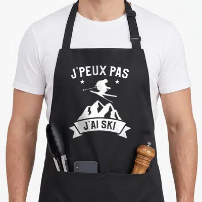 Tablier de cuisine noir avec silhouette de skieur, montagnes et texte J'peux pas j'ai ski en blanc.