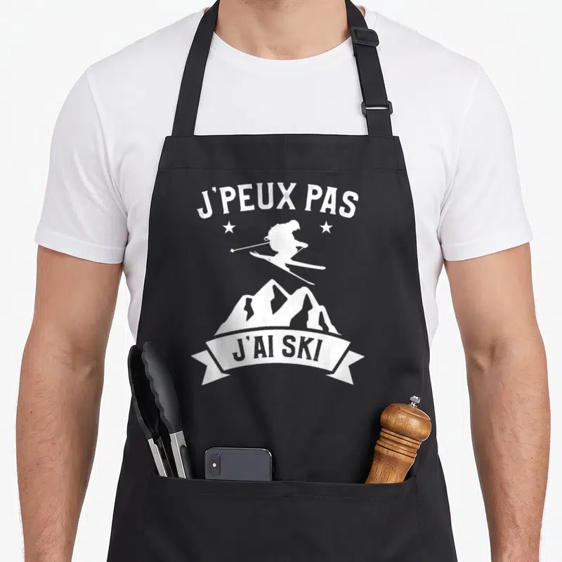 Tablier de cuisine noir avec silhouette de skieur, montagnes et texte J'peux pas j'ai ski en blanc.