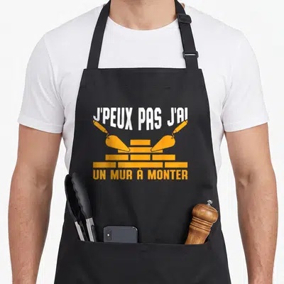 Tablier de cuisine noir avec inscription j'peux pas j'ai un mur à monter et deux truelles orange posées sur des briques.