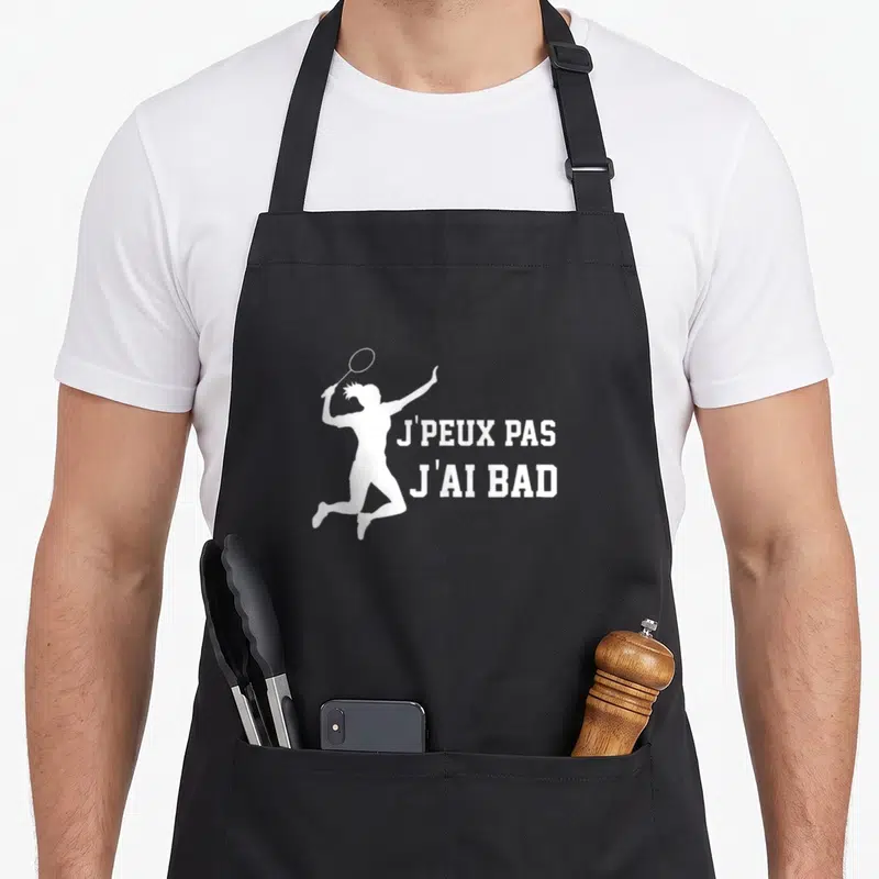 Tablier de cuisine noir avec silhouette blanche d'un joueur de badminton en saut tenant une raquette et le texte j'peux pas j'ai bad.