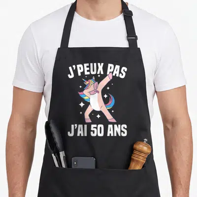 Tablier de cuisine noir avec une licorne rose effectuant un dab entourée d'étoiles et du texte blanc J'peux pas j'ai 50 ans