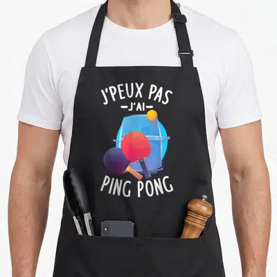 Tablier de cuisine noir avec texte J'peux pas j'ai Ping Pong et illustration de raquettes colorées devant une table.