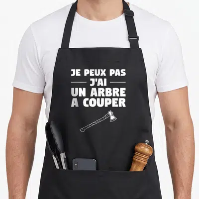 Tablier de cuisine noir avec l'inscription je peux pas j'ai un arbre à couper et une hache blanche en dessous.
