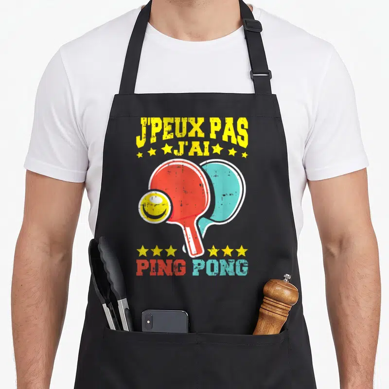 Tablier de cuisine noir avec illustration de deux paddles ping pong et texte jpeux pas jai ping pong en lettres colorées.