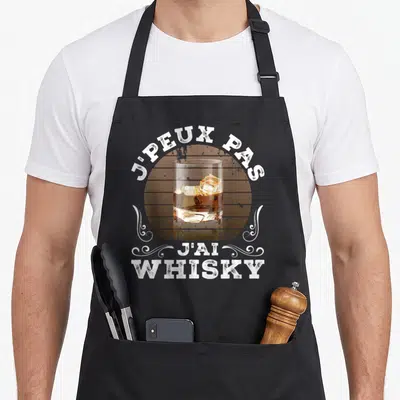 Tablier de cuisine noir avec un verre de whisky sur fond bois et texte blanc en arc de cercle.