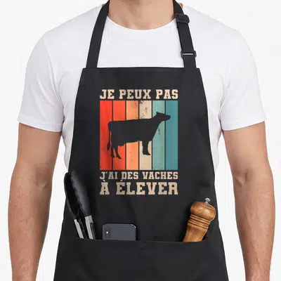 Tablier de cuisine noir avec silhouette de vache sur fond de bandes colorées et texte beige Je peux pas j'ai des vaches à élever