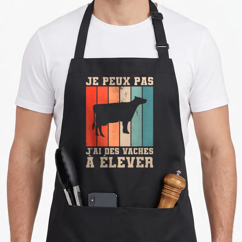 Tablier de cuisine noir avec silhouette de vache sur fond de bandes colorées et texte beige Je peux pas j'ai des vaches à élever
