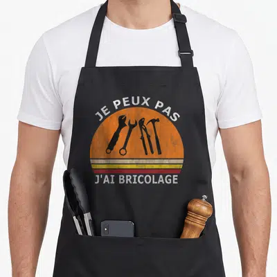 Tablier de cuisine noir avec motif circulaire orange affichant des outils et le texte je peux pas j'ai bricolage.