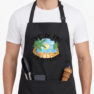 Tablier de cuisine noir avec illustration de pieds nus sur le sable face à l'océan et palmiers encadrant le texte.