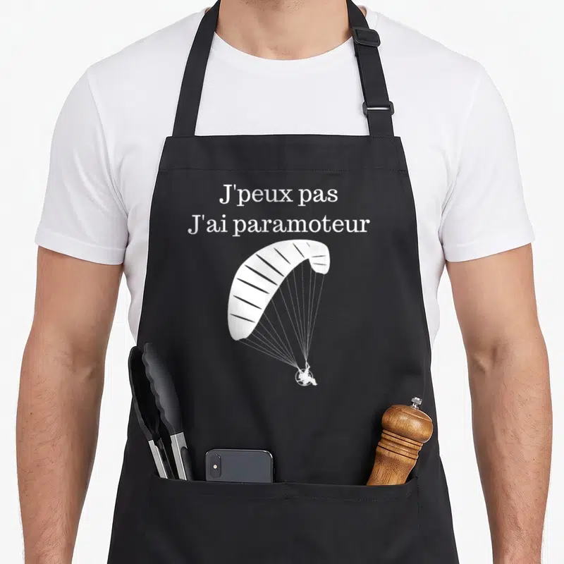 Tablier de cuisine noir avec l'inscription J'peux pas J'ai paramoteur et un dessin de voile blanche.