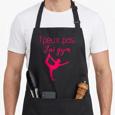 Tablier de cuisine noir avec une silhouette de gymnaste rose et le texte j'peux pas j'ai gym imprimé en haut.