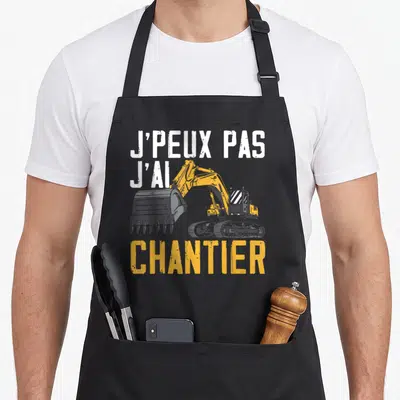 Tablier de cuisine noir avec texte blanc et excavateur jaune illustrant l'expression j'ai chantier.
