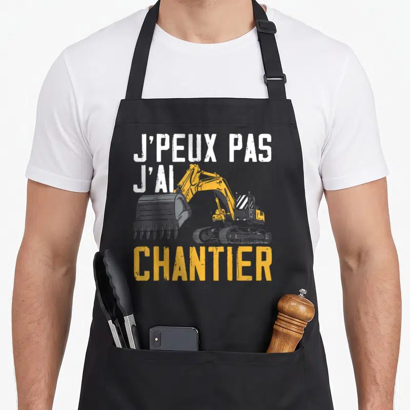 Tablier de cuisine noir avec texte blanc et excavateur jaune illustrant l'expression j'ai chantier.