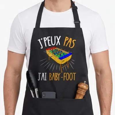 Tablier de cuisine noir avec illustration colorée d'une table de baby-foot et texte blanc et orange.