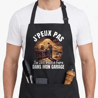 Tablier de cuisine noir avec une illustration circulaire d'un homme travaillant dans un garage rempli d'outils et le texte j'peux pas j'ai des trucs à faire dans mon garage.
