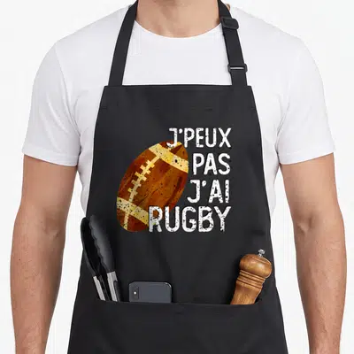 Tablier de cuisine noir avec illustration de ballon de rugby et phrase J'peux pas j'ai rugby en lettres blanches texturées.