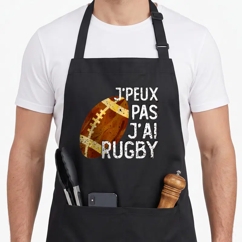 Tablier de cuisine noir avec illustration de ballon de rugby et phrase J'peux pas j'ai rugby en lettres blanches texturées.