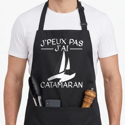 Tablier de cuisine noir avec illustration blanche d'un catamaran et le texte J'peux pas j'ai catamaran imprimé au centre.