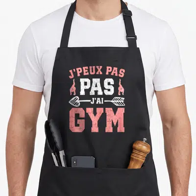 Tablier de cuisine noir avec inscription j'peux pas j'ai gym en rose et blanc accompagnée de silhouettes de gymnastes et d'une flèche.