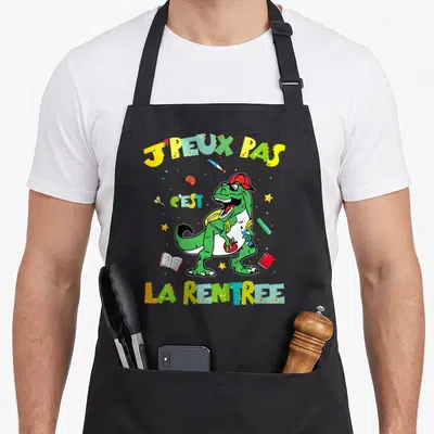 Tablier de cuisine noir avec illustration colorée d'un dinosaure portant un sac à dos et tenant un crayon sous le texte J'peux pas c'est la rentrée.