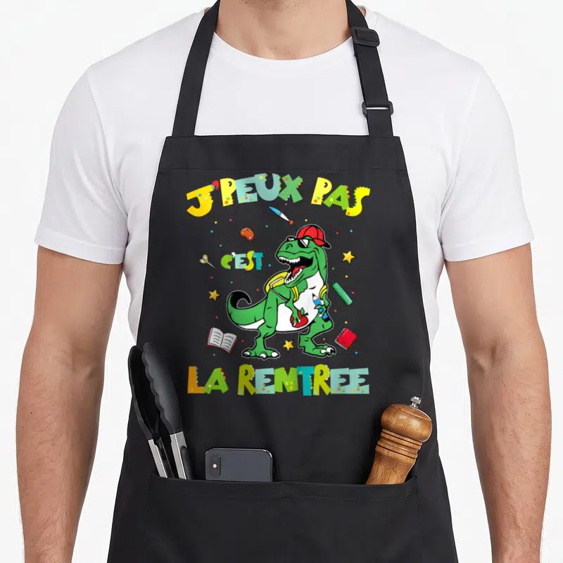Tablier de cuisine noir avec illustration colorée d'un dinosaure portant un sac à dos et tenant un crayon sous le texte J'peux pas c'est la rentrée.