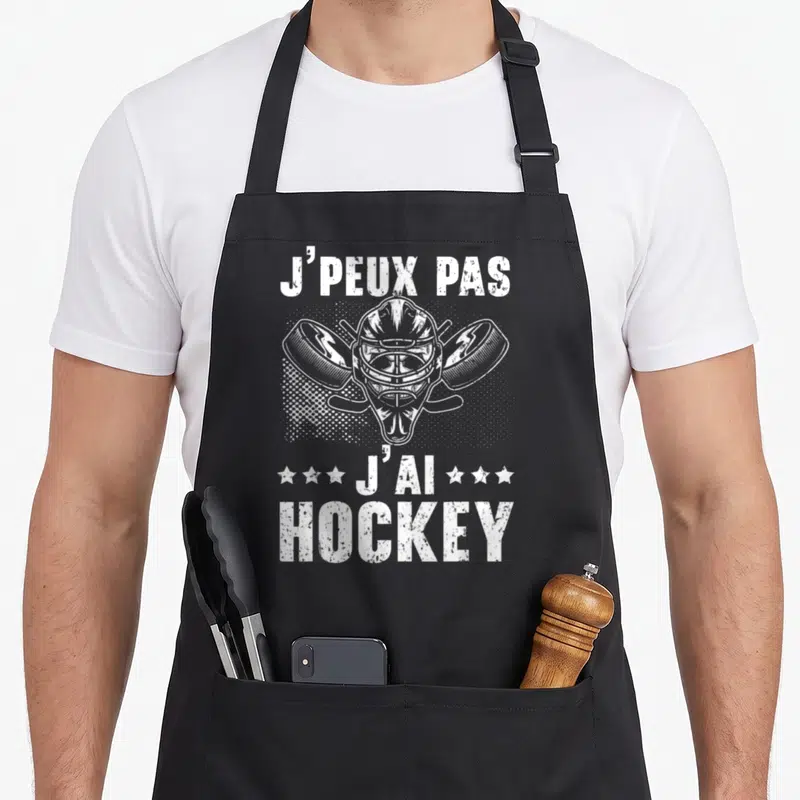 Tablier de cuisine noir avec illustration de masque de gardien de hockey, deux crosses et des disques en blanc sur fond sombre.