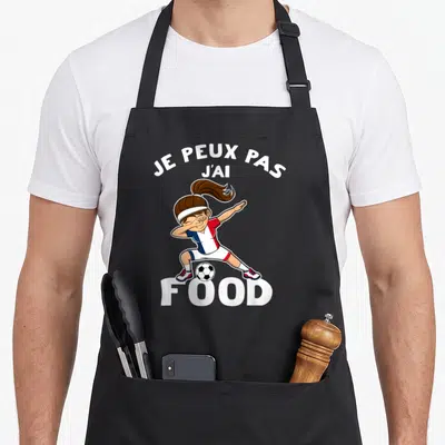 Tablier de cuisine noir avec une footballeuse faisant un dab au-dessus d'un ballon de football.