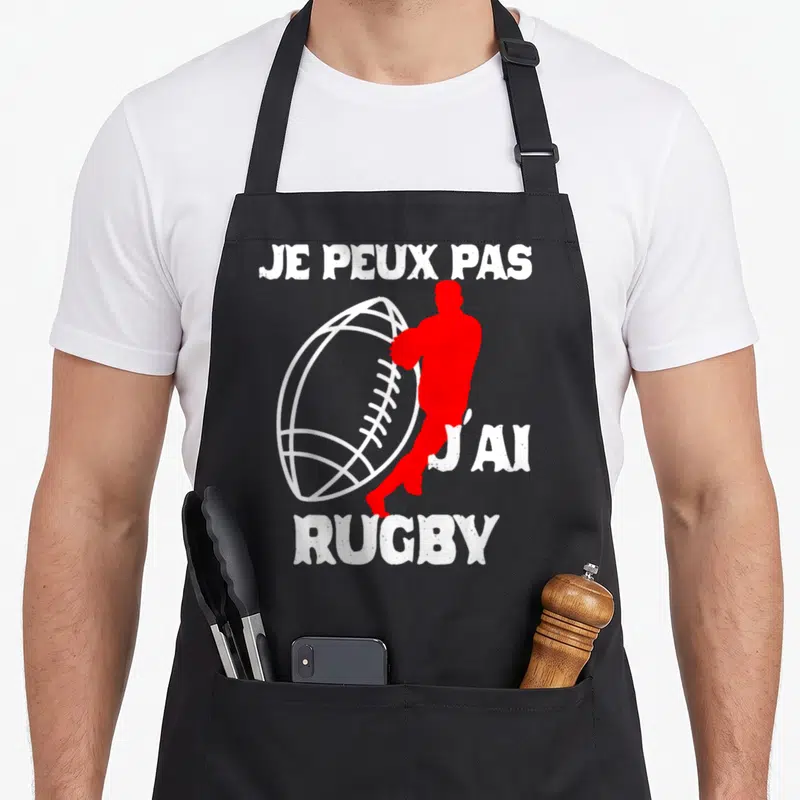 Tablier de cuisine noir avec silhouette rouge tenant un ballon de rugby et texte blanc humoristique.