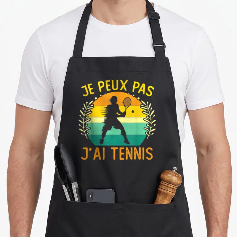 Tablier de cuisine noir avec silhouette de joueur de tennis et texte je peux pas j'ai tennis sur fond de soleil coloré.