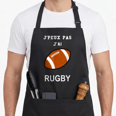 Tablier de cuisine noir avec une balle de rugby orange et le texte j'peux pas j'ai rugby en blanc.