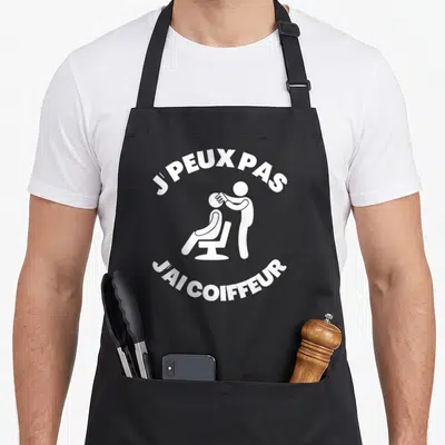 Tablier de cuisine noir avec inscription j'peux pas j'ai coiffeur et silhouette blanche d'un coiffeur au travail.