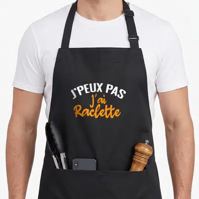 Tablier de cuisine noir avec l'inscription j'peux pas j'ai raclette en blanc et orange.