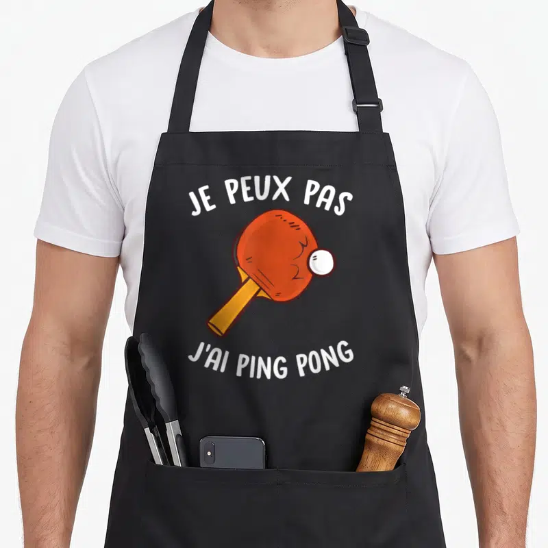 Tablier de cuisine noir avec une raquette de ping-pong orange, un ballon blanc et le texte je peux pas j'ai ping pong en blanc.
