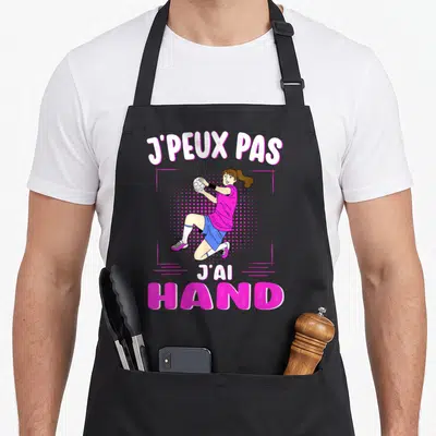 Tablier de cuisine noir avec une femme en maillot rose et short bleu tenant un ballon de handball sous le texte j'peux pas j'ai hand.