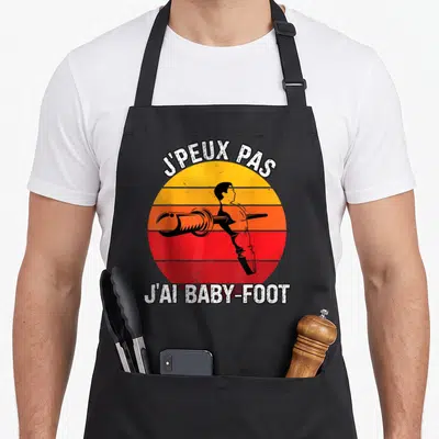 Tablier de cuisine noir avec motif rétro JPEUX PAS JAI BABY-FOOT et silhouette stylisée sur fond dégradé.