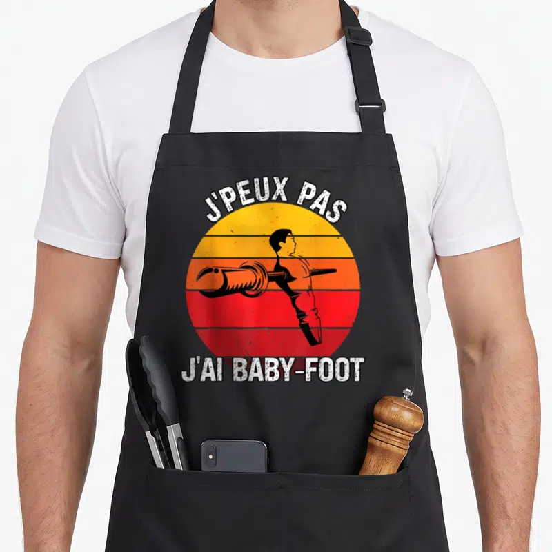 Tablier de cuisine noir avec motif rétro JPEUX PAS JAI BABY-FOOT et silhouette stylisée sur fond dégradé.