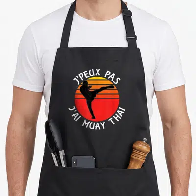 Tablier de cuisine noir avec silhouette boxeuse et texte j'peux pas j'ai muay thai sur fond de soleil rétro.