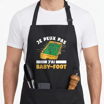 Tablier de cuisine noir avec illustration d'une table de baby-foot jaune et le texte je peux pas j'ai baby-foot.