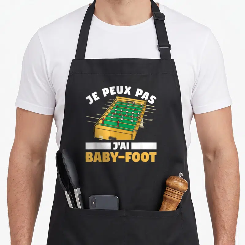 Tablier de cuisine noir avec illustration d'une table de baby-foot jaune et le texte je peux pas j'ai baby-foot.